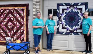 Three members of the Tracy CCW (Tracy, MN) stand in front of a garage which displays two beautiful geometric quilts- one blue and white, and one burgundy and white.